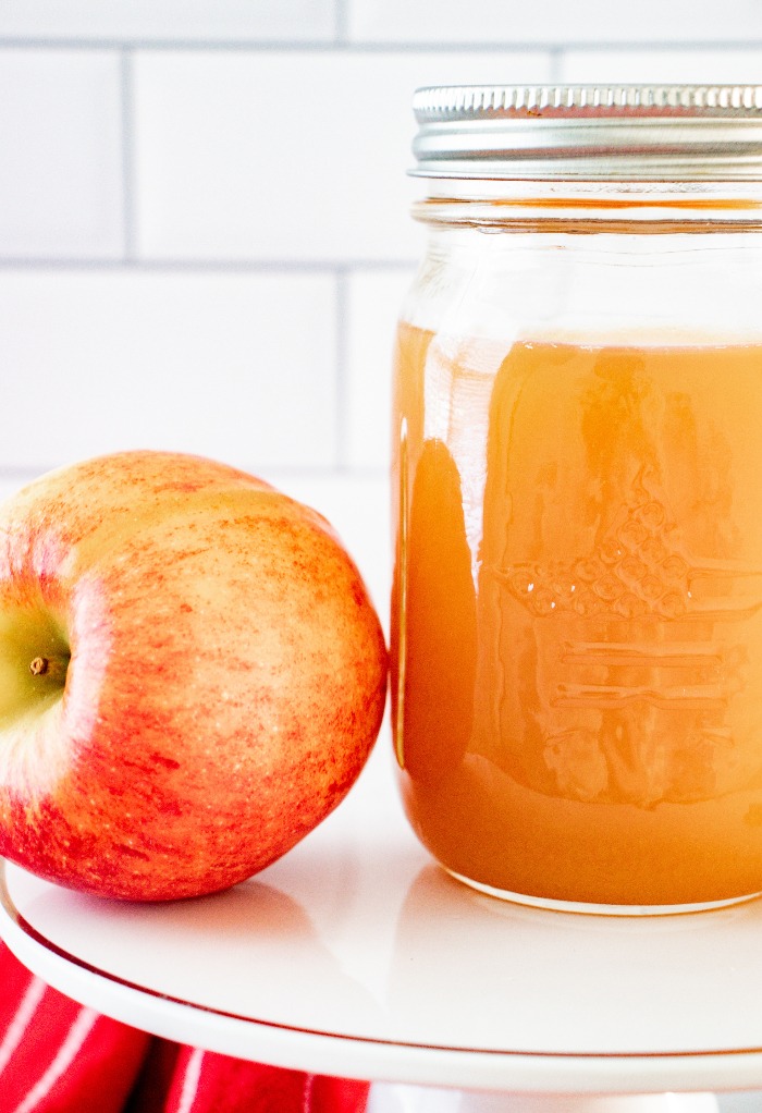 A clear mason jar with homemade apple pie moonshine on a white cake plate with a fresh apple on the side.