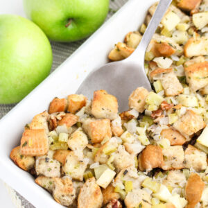 Scooping up with a silver spoon apple stuffing in a white baking dish.