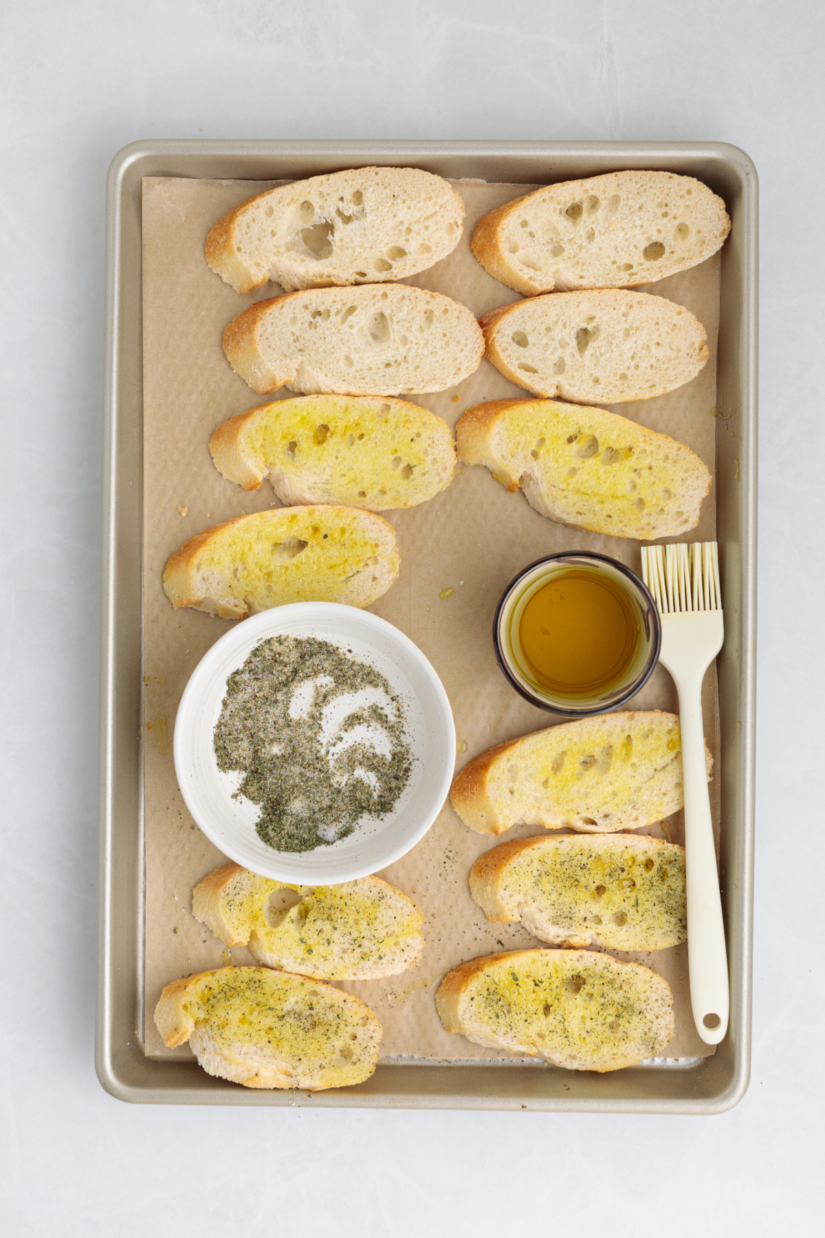 Sliced baguette slices on a baking sheet with olive oil and seasoning before toasting in the oven.