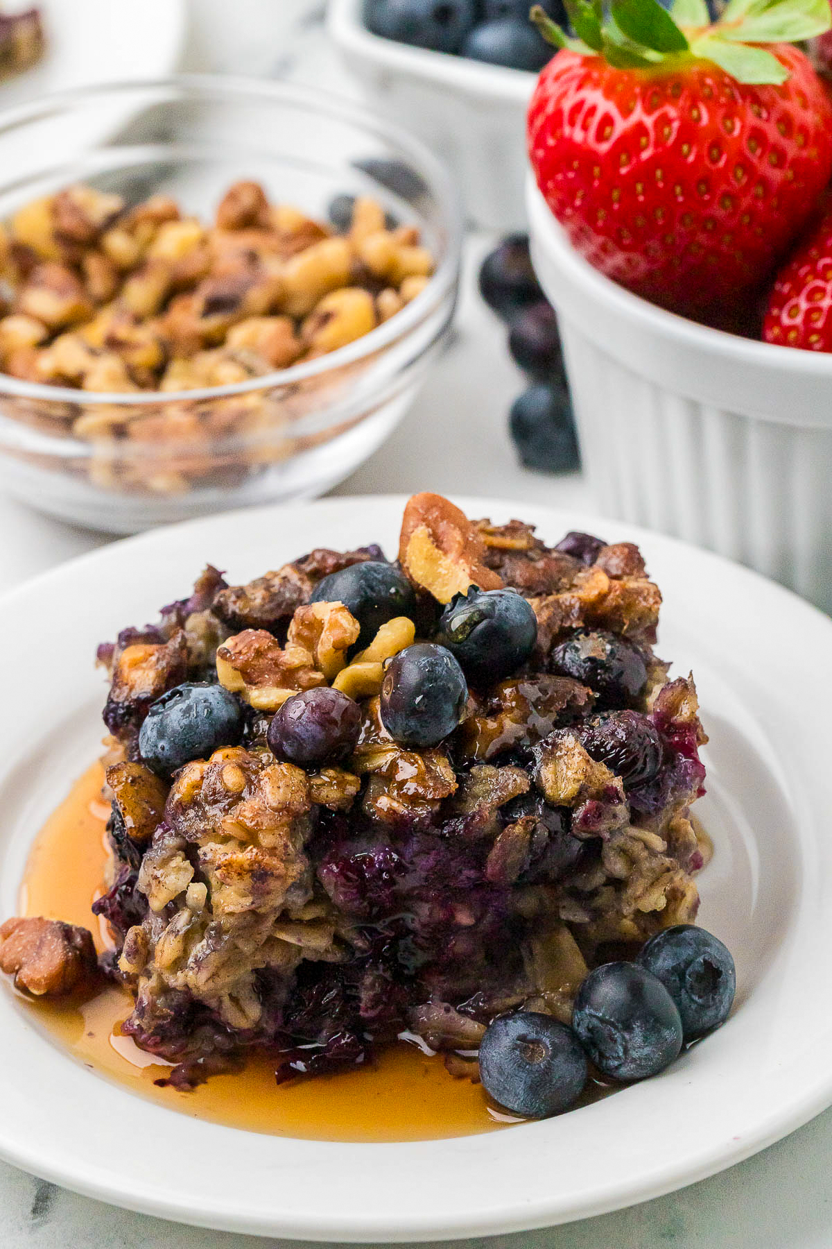 A slice of blueberry baked oatmeal topped with maple syrup on a white serving plate with fresh strawberries on the side.
