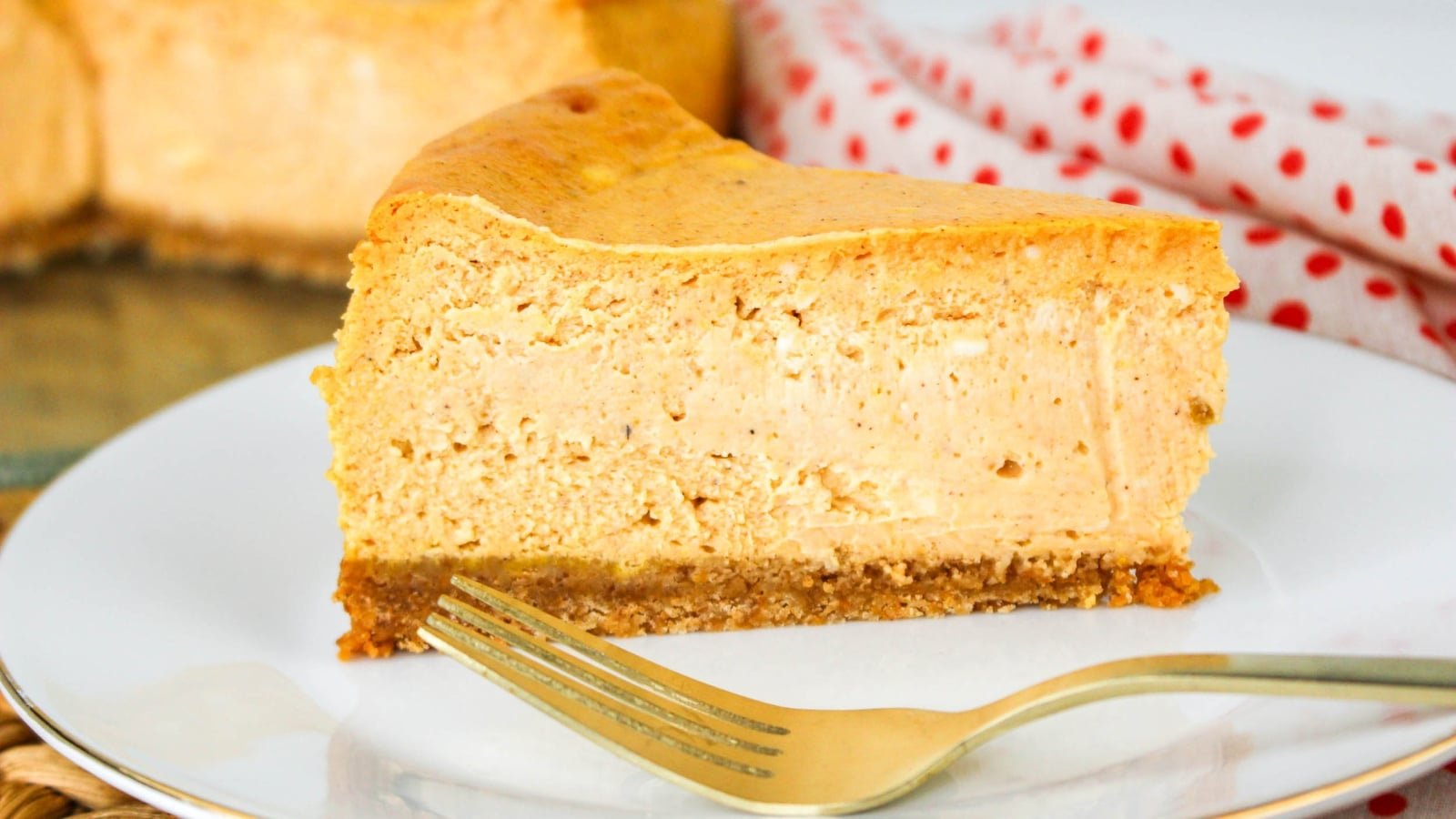 A slice of cheesecake on a plate with a silver fork, in front of a white cloth with red dots.