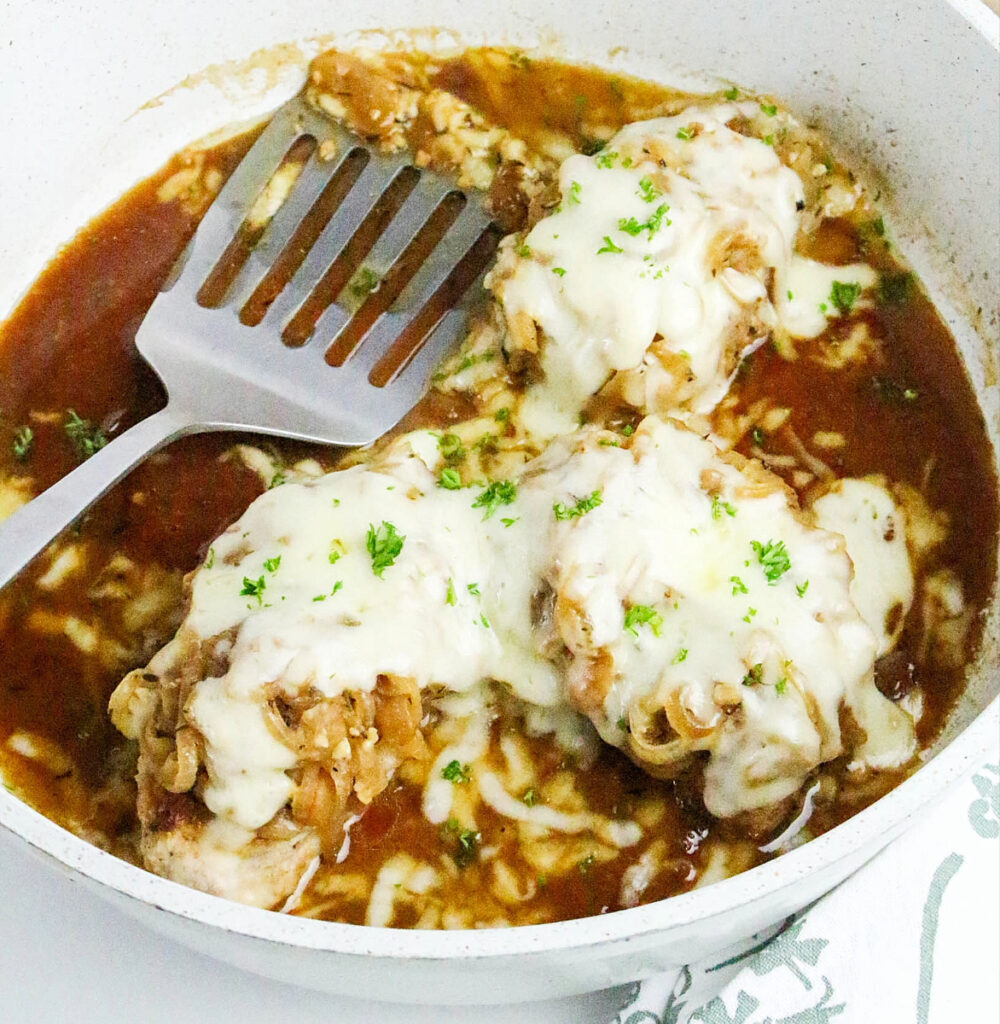French onion pork chops in a white skillet topped with melted cheese.