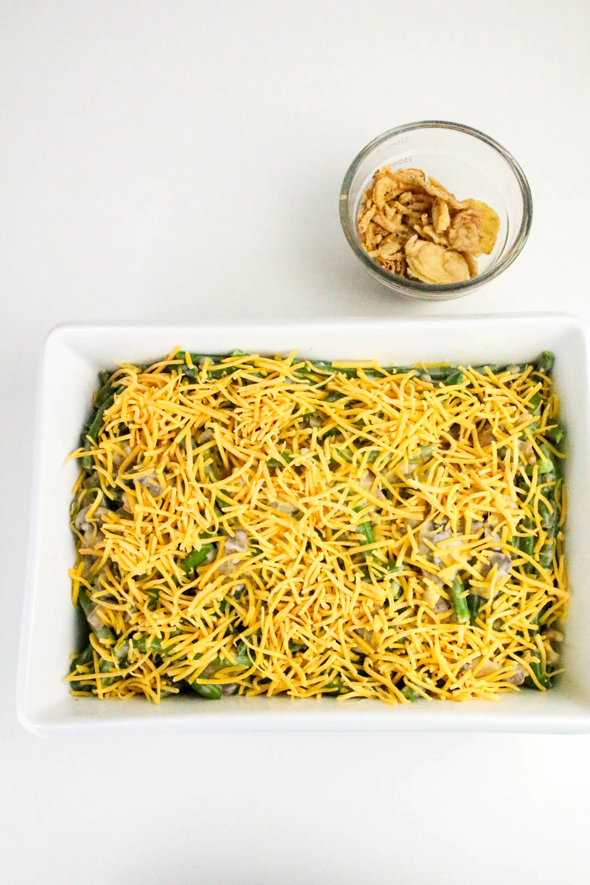 Green beans casserole with fresh green beans topped with shredded cheddar cheese in a white baking dish.
