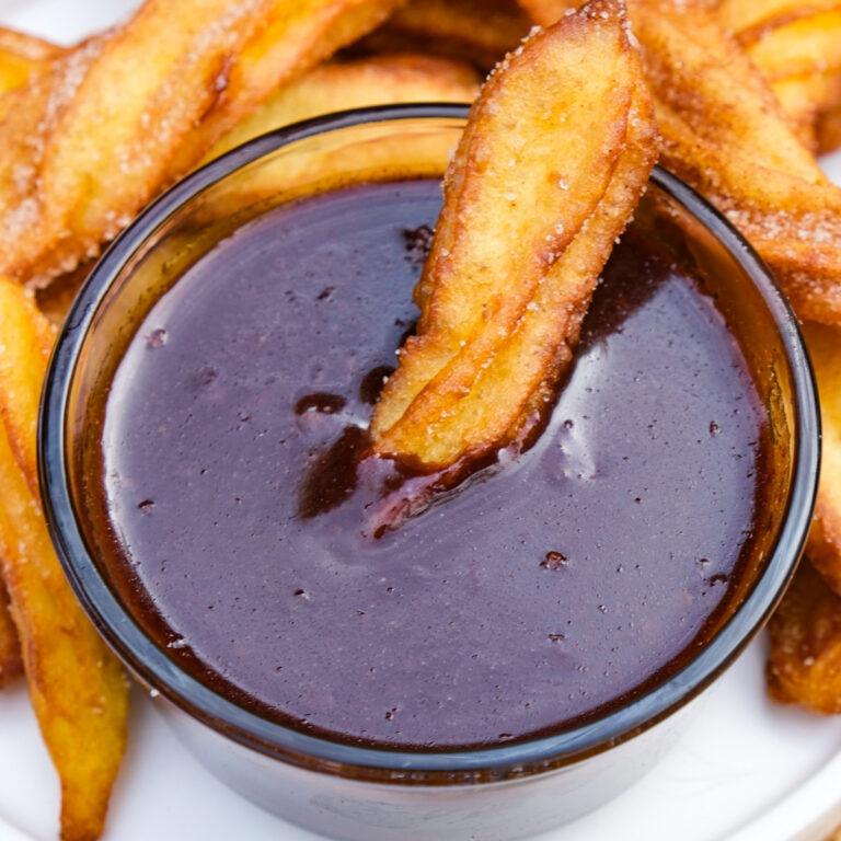 Homemade Ghirardelli chocolate sauce in a small clear serving bowl with a churro dipped in the center.