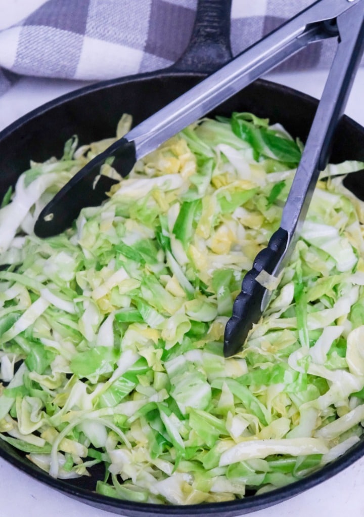 Sautรฉing cabbage in a cast iron skillet with tongs.