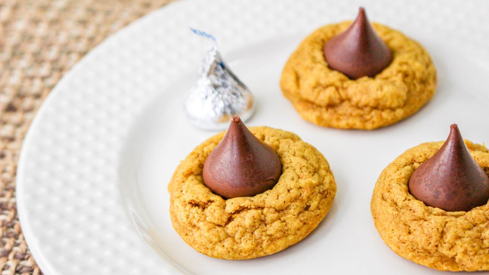 Three peanut butter cookies topped with chocolate kisses are arranged on a white plate, next to an unwrapped chocolate kiss.