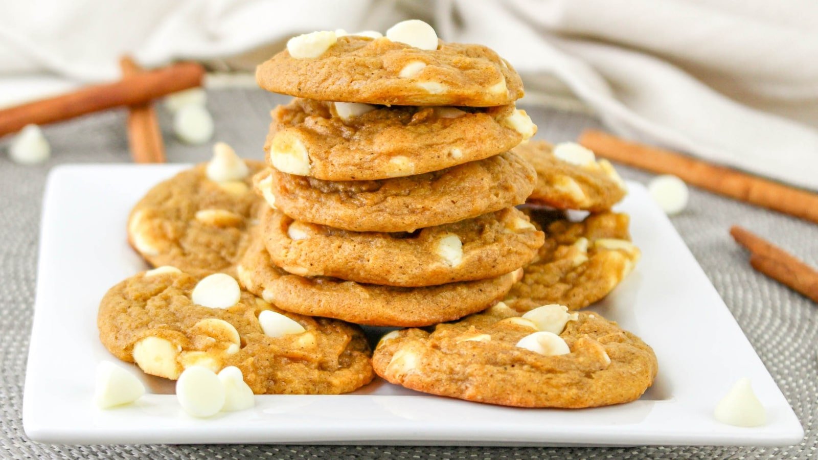 A stack of cookies with white chocolate chips on a white plate, surrounded by more chips and cinnamon sticks.
